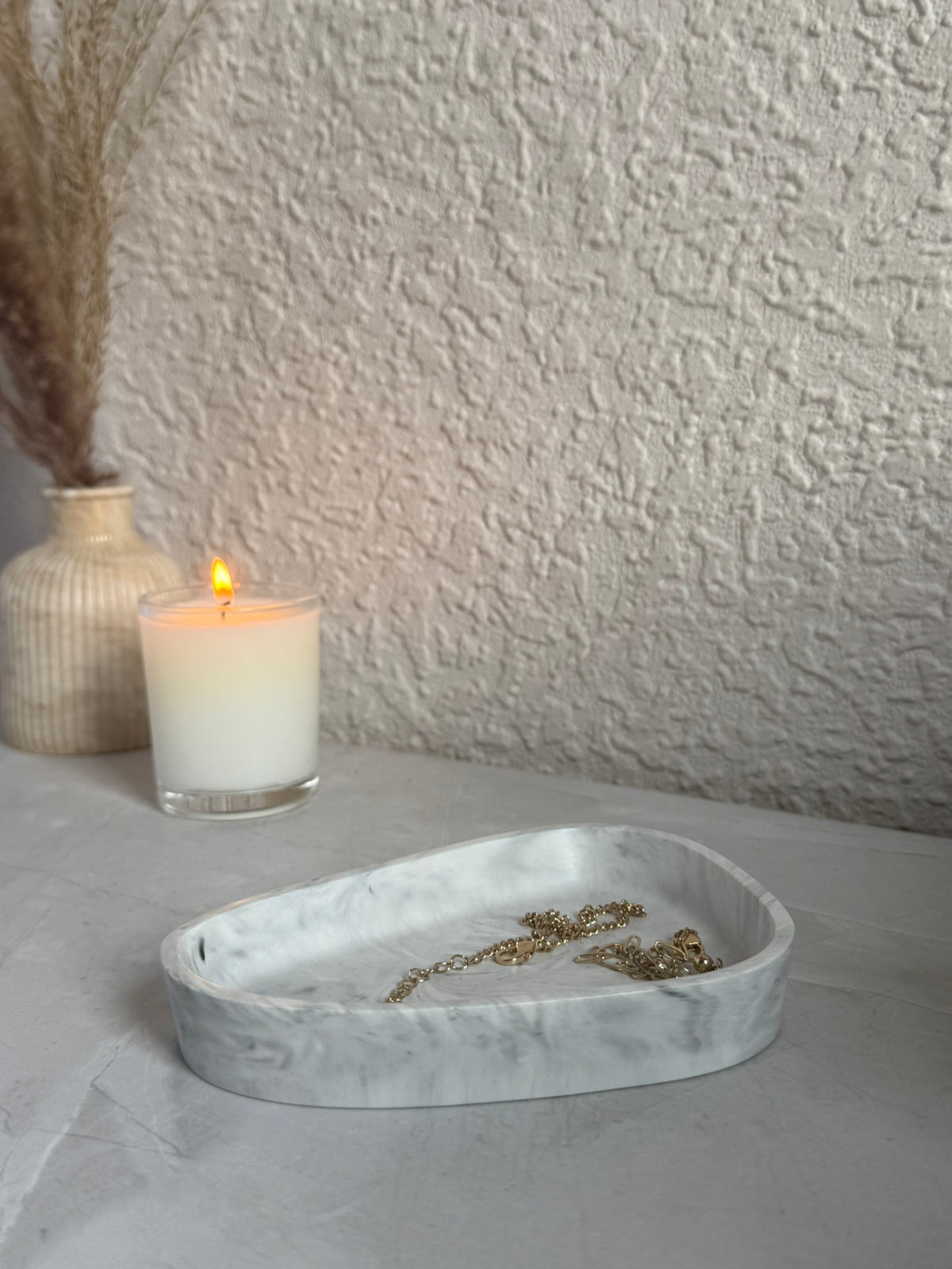 A white marble-patterned oval jewelry tray holding gold-toned jewelry, styled on a light surface with a lit white candle and a textured beige wall in the background.