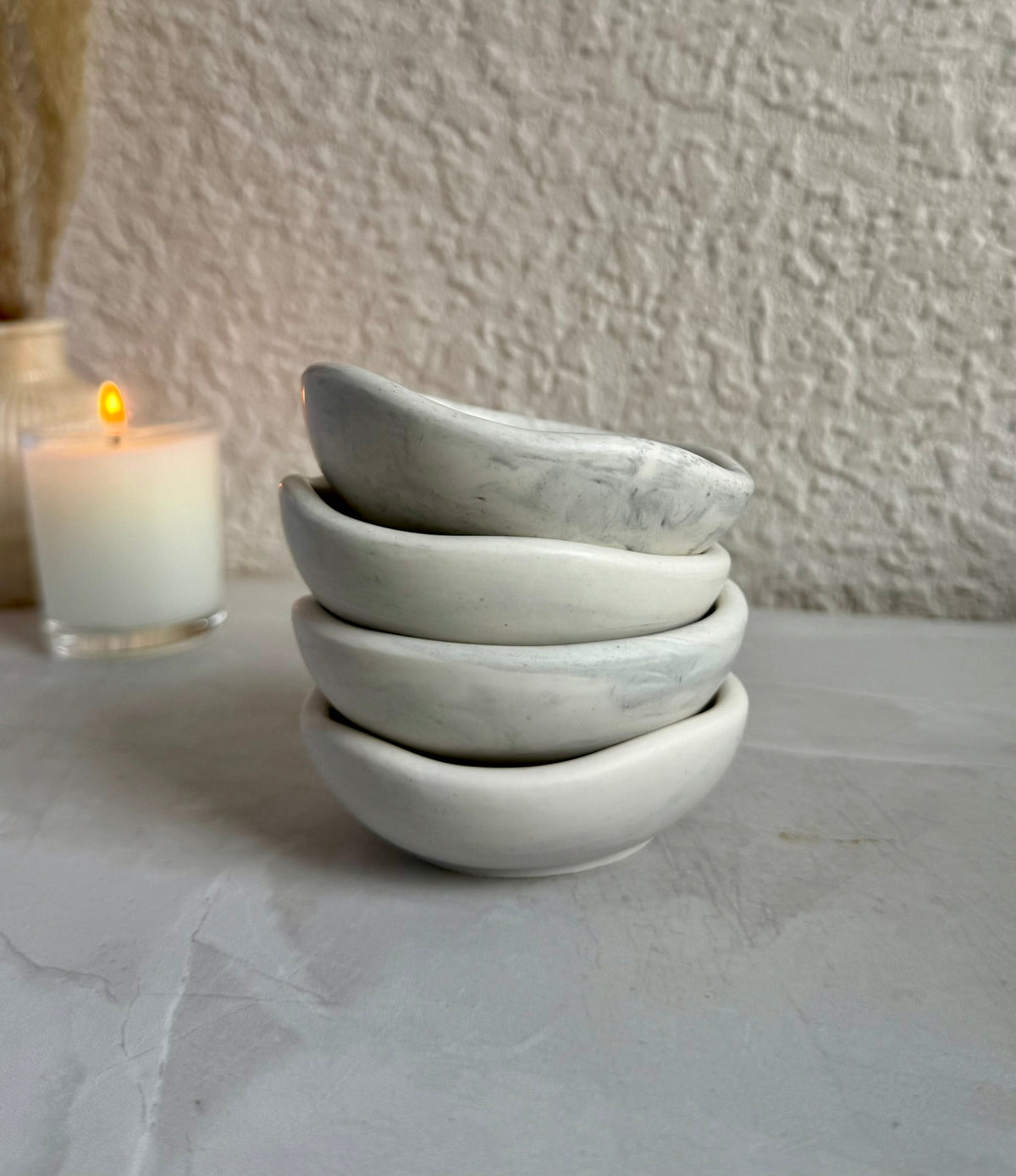 Four small stacked ceramic bowls with a white and gray marble pattern, set on a light gray surface with a lit candle in the background.